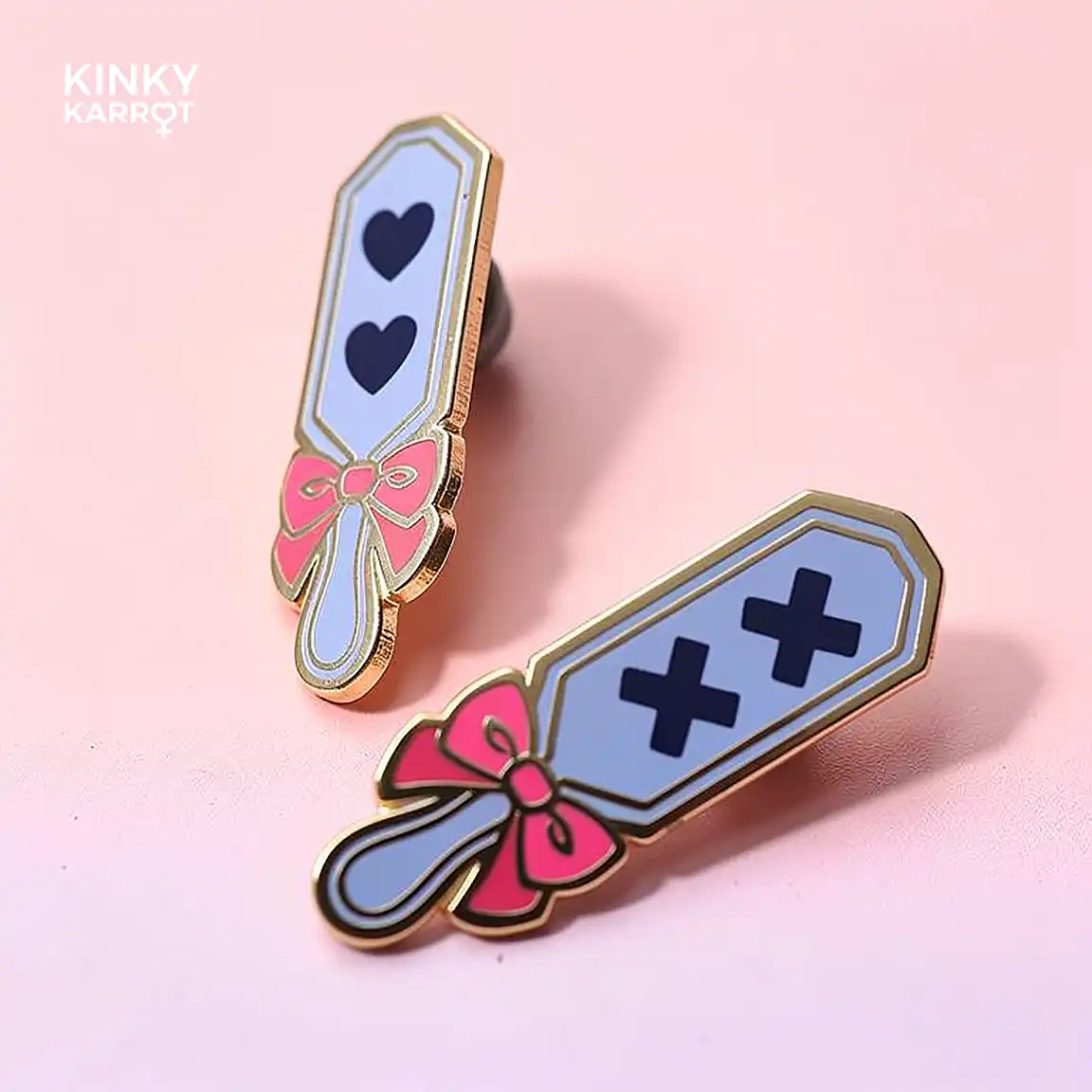 Two pastel Spanking Paddle Enamel Pin with different designs – one with black hearts, one with black Xs – lying on a pink background.