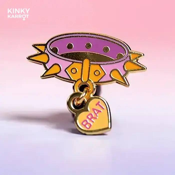Pink spiked collar enamel pin with “BRAT” charm on pastel background