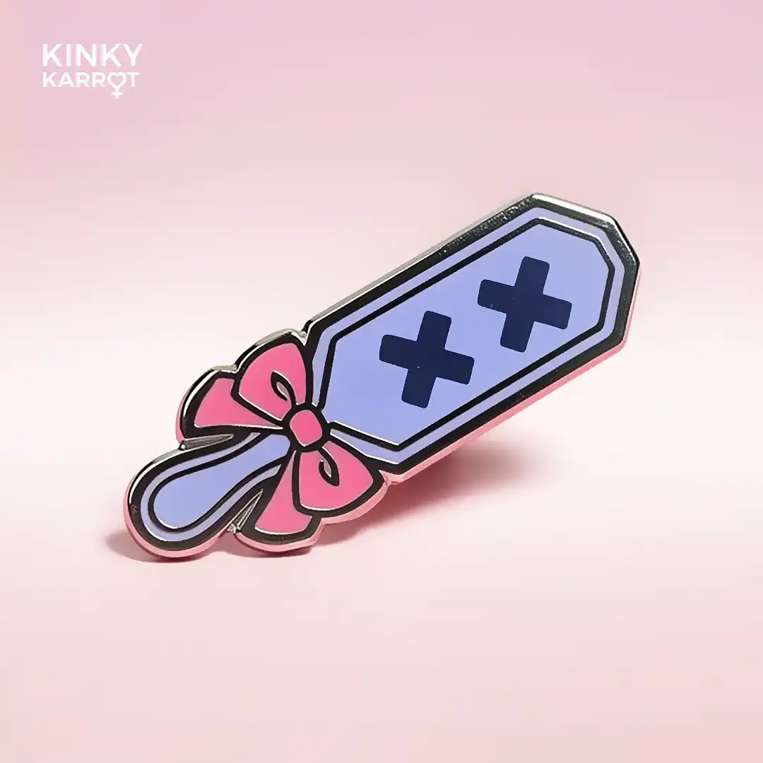 Single pastel spanking paddle enamel pin with pink bow and two black X marks on a lavender surface.